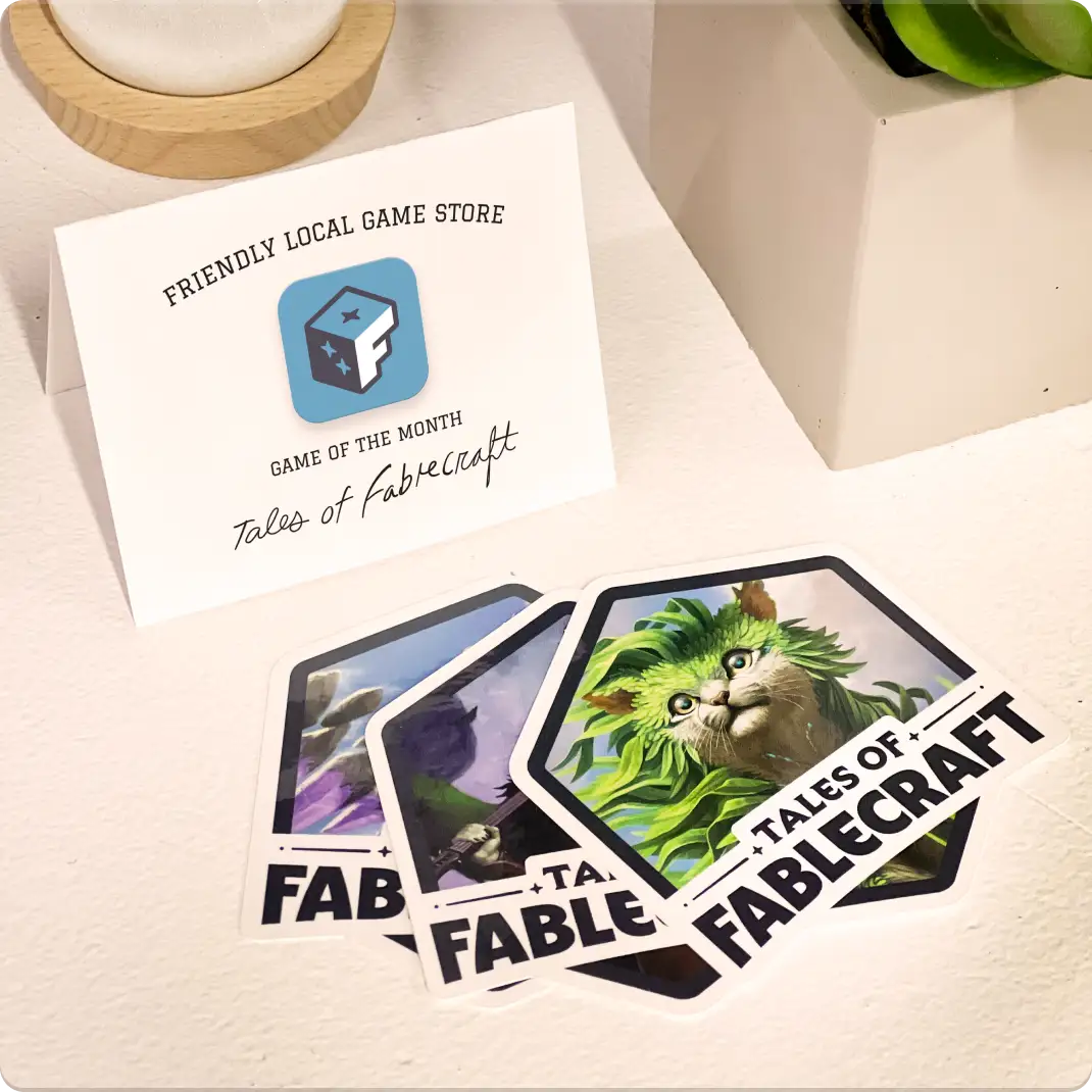 A counter at a local game store holds a printed sign that reads: Game of the Month: Tales of Fablecraft. Three Fablecraft stickers are fanned out beside it.